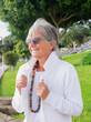 © luciano - Smiling senior woman in public park having relaxed moments and looking away. Mature gray haired woman white dressed with necklace and sunglasses enjoying retired lifestyle in outdoor