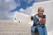 © luciano - Happy adult mature senior woman sitting outdoors on staircases using smartphone smiling. Active elderly female denim dressed relaxing enjoying tech and social
