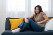 © Prot - Woman Abdominal Discomfort: A woman sits on a sofa, clutching her abdomen with a pained expression, indicating internal discomfort and highlighting the human experience of physical struggle.
