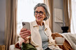 © HBS - Portrait of a beautiful smiling middle-aged gray-haired woman wearing glasses, using a phone while sitting in a cozy living room with a cup of coffee or tea.