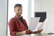 © Prostock-studio - A man sits at a desk in an office. He holds two sheets of paper and looks at them with a smile. Sunlight comes through the window, creating a bright atmosphere.