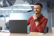 © Prostock-studio - A man is seated at a desk inside an office, looking at a laptop screen. He appears engaged and is smiling while taking part in an online meeting. The office has modern decor.