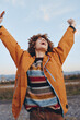 © SHOTPRIME STUDIO - Happy woman with curly hair wearing a rainbow sweater and orange jacket raises arms outdoors. Woman smiles joyfully holding smartphone, lifestyle concept with natural light and open sky background.