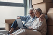 © Daniel - An affectionate senior couple sits on the floor among moving boxes, smiling and using a laptop as they unpack and settle into their new home, sharing a relaxed, joyful moment together.