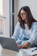 © Daniel - Young woman sits at a table by a window, smiling as she types on a laptop. Casual remote work setup with notebook, coffee cup and natural light conveying focus, comfort and productivity.