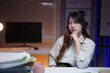 © MINAE - Young Asian woman feeling tired and stressed, sitting at her office desk with a pile of documents, working late into the night and experiencing burnout from overwork