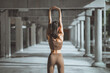 © oatawa - Back view of a fit young woman in brown activewear stretching her arms overhead to improve flexibility during a morning workout in a modern industrial concrete hallway, focusing on wellness.
