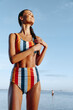 © SHOTPRIME STUDIO - Young woman in colorful striped swimsuit stands by the sea, eyes closed, enjoying sunlight and fresh breeze, wet skin glistening, relaxed lifestyle moment outdoors under clear blue sky