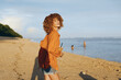 © SHOTPRIME STUDIO - A young woman with curly hair wearing an orange shirt and denim shorts stands on a sandy beach holding a drink, enjoying a sunny day by the calm sea. Woman lifestyle concept.