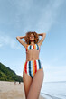 © SHOTPRIME STUDIO - Young woman with curly hair wearing colorful striped bikini standing on the beach with hands behind head. Sunny day with blue sky and calm sea in background. Relaxed lifestyle concept.