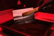 © Stitch - Hand wearing a black glove using bamboo tongs to submerge a photograph in developer fluid under red darkroom light