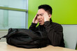 © fotolite - A young Asian male student holds his temples with both hands in frustration at a desk with a backpack in front, ideal for academic stress, student burnout, or mental health awareness concepts.