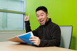 © fotolite - A young Asian male student raises his index finger with a big smile while holding a notebook at a desk, ideal for eureka moment, creative thinking, or student success concepts.