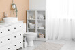 © Pixel-Shot - Interior of restroom with toilet bowl, chest of drawers and shelving unit near beige wall