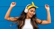 © Tiaga - Cute tanned woman with long brown hair wearing white top and yellow bandana listens to music in wireless headphones and dances on blue wall background.