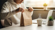 © Pandu - Man taking a lunch break while working from home opening a food delivery bag at his desk with a laptop and coffee
