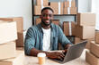 © Ranazain - A joyful African American man smiles working on his laptop surrounded by many cardboard shipping boxes in his successful home office for a thriving e