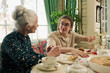 © TrueFrame Collective - Senior Caucasian woman talking with teenage Caucasian boy sitting at table, teenager smiling and adding jam to pastry, both engaging in conversation during breakfast or tea time
