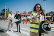 © qunica.com - Woman engineer in a hi vis vest speaks into a walkie talkie while holding a hard hat and tablet. Two colleagues consult blueprints in the blurred background on a road construction site.