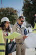 © qunica.com - A construction team of engineers and an architect review blueprints at an outdoor job site. The professionals consult plans and documents while wearing safety vests and a hard hat.