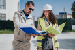 © qunica.com - Two construction professionals review project documents on an outdoor building site. Male and female engineers consult plans while wearing hard hats and high-visibility vests.