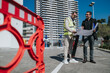 © qunica.com - Two people, a construction engineer in a safety vest and an architect, review large building plans outdoors. They stand on-site discussing blueprints in front of a high-rise under a clear blue sky.