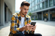 © Vadym - Young man in checkered shirt with wireless earbuds looks at smartphone. Sits outdoors near modern office buildings. Uses mobile device, browses content. Guy appears focused, engaged with phone.