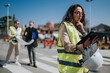 © qunica.com - Female construction engineer in a high-visibility vest reviews plans on a tablet. Two colleagues discuss blueprints in the background at a roadwork site.