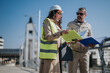 © qunica.com - A female engineer in a high-visibility vest and hardhat consults plans with a male colleague outdoors. The pair review project documents during a site inspection near an urban bridge.