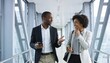© SERHII - Two professional black colleagues walking and talking in a modern office corridor. Businessman and businesswoman networking and collaborating on a project.