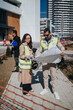 © qunica.com - Male and female architects in high-visibility vests examine blueprints at an outdoor urban construction site. They discuss plans and check documents during an on-site meeting near modern buildings.