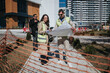 © qunica.com - Two construction professionals in high-visibility vests examine blueprints and digital notes on site. Colleagues review project plans beside orange safety fencing at a new urban development.