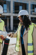 © qunica.com - A female construction engineer wearing a hard hat and high-visibility vest reviews plans on a building site. She discusses the blueprints with a colleague while overseeing construction progress.