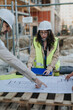 © qunica.com - A female construction engineer in a hard hat and high-visibility vest reviews blueprints at a job site. Colleagues point at plans on a pallet while they discuss site layout and project details.