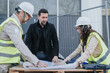 © qunica.com - Construction engineers review blueprints and plans at an outdoor site while wearing safety gear. The team discusses project details over plans on a makeshift table at the construction site.