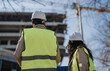 © qunica.com - Two construction engineers wearing hard hats and high-visibility vests inspect a building site from the rear. A back-view scene showing professionals collaborating on an urban development project.