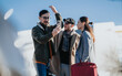 © qunica.com - Three friends pose for a selfie while holding luggage and wearing winter coats. They stand together in an urban setting with clear blue sky and soft sunlight.
