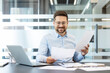 © Liubomir - Smiling businessman reviewing business documents, financial reports, and contracts while working at a desk with a laptop in a modern office, planning strategies for success