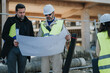 © qunica.com - Construction engineers study a large blueprint while standing at an active building site. Two professionals in hard hats and high-visibility vests discuss project plans on site.