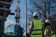 © qunica.com - Two construction engineers wearing hard hats and high-visibility vests inspect an urban building site. The colleagues assess progress and site conditions near a crane and partially built structure.