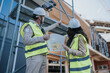 © qunica.com - Two construction engineers in hard hats and high-visibility vests review a site blueprint. They stand by scaffolding at a building under construction discussing plans.