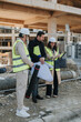 © qunica.com - Three construction engineers wearing hard hats and high-visibility vests review blueprints and a tablet on site. They stand in front of an unfinished concrete building discussing plans and inspection