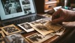 © Afsana - An elderly person's hand reaches for a faded vintage photograph among many scattered pictures on a dark wooden desk next to a modern open laptop