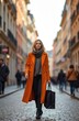 © miss irine - Blonde woman walks down European city street wearing orange trench coat, carrying reusable shopping bag. Looks fashionable, confident during autumn weather. Woman enjoys urban exploration, city walk.