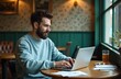 © miss irine - Bearded man sits at table in cafe working on laptop. He wears a casual sweater and types on keyboard while drinking beverage. Focused male person uses notebook for remote job.