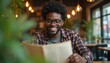 © Vadym - Smiling African American man wearing glasses reads menu in cafe. Blurred background with plants and bokeh lights. He enjoys picking food and drink for meal.