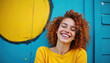 © Vadym - Young woman with red curly hair laughs joyfully against a bright blue wall with yellow circle art. She wears a yellow shirt and has freckles, conveying a feeling of pure happiness.