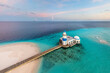 © AmazingAerialAgency - Aerial view of a vibrant turquoise sea contrasting with the deep blue, surrounding a pristine white sandy beach and a striking white and blue mosque connected by a wooden pier, Maldives, Maldives.