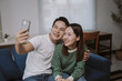 © Crystal - Young Asian couple smiling, making a selfie together using a smartphone while relaxing on the couch in their living room