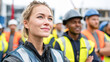 © Kowit - A confident female construction worker wearing safety gear stands with her team in a worksite, symbolizing diversity and teamwork in the industry.